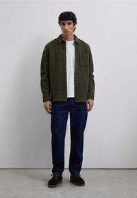 Camisa chaqueta de mezcla de lana verde con botón en el frente, bolsillo en el pecho; combinada con pantalones de mezclilla azul oscuro y zapatos de cordones marrón oscuro.