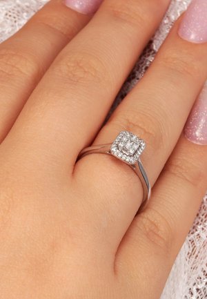 Eine Hand mit hellpinkglänzenden Nägeln trägt einen silbernen Ring, der auf dem Ringfinger einen quadratischen Cluster aus kleinen funkelnden Diamanten zeigt.