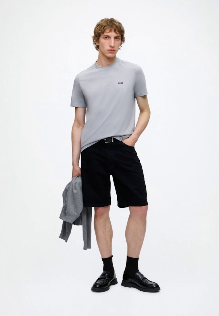 Jeune homme portant un T-shirt gris clair, un short noir, des chaussettes noires et des chaussures noires, tenant un sweat-shirt gris dans une main.
