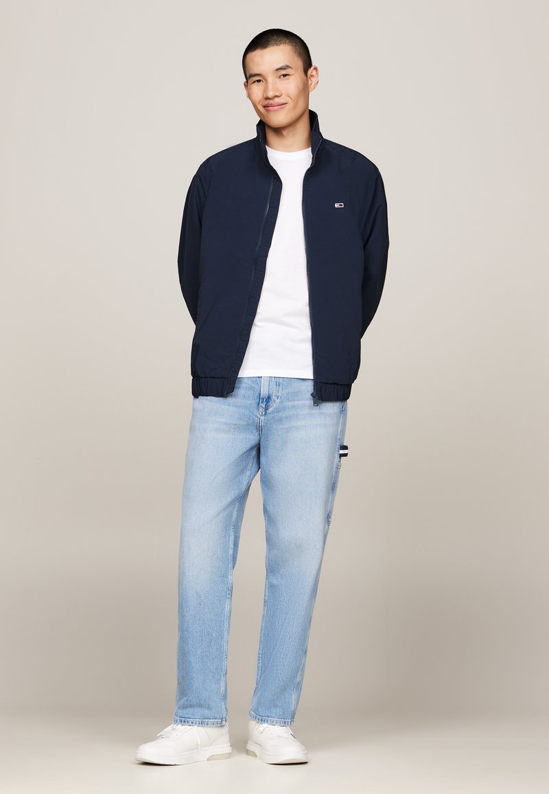 Veste de pilote marine avec une fermeture éclair à l'avant, des poignets élastiques et un petit logo, associée à un jeans bleu clair et des baskets blanches. Design simple.