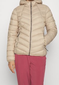 Beige puffer jakke med hætte, der har chevron syninger, lynlås lukning og sidelommer. Bæres med pink bukser.
