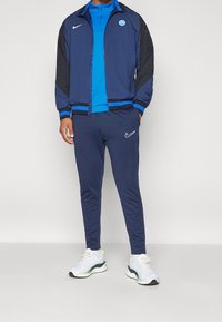 Navy blue track jacket with black sleeves and blue accents, featuring a logo. Matched with navy pants and white athletic shoes.