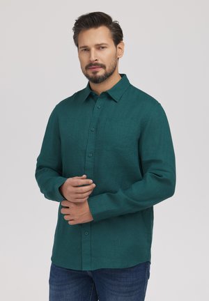 Mężczyzna z krótkimi włosami i brodą, ubrany w ciemnozieloną koszulę na guziki i niebieskie dżinsy, stojący na tle jednolitego jasnego tła.