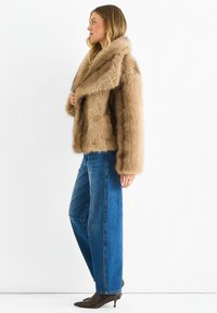 Faux fur brown jacket with a wide collar, soft texture, and oversized shape, paired with high-waisted blue jeans and dark heeled shoes.