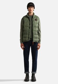 Grüne gepolsterte Weste über einem grauen Pullover, dunkelblauen Jeans und schwarzen Stiefeln. Die Weste hat ein rundes Patch und die Jeans haben umgeschlagene Bündchen.