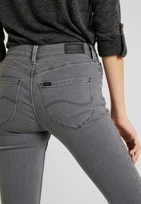 Jeans de mezclilla gris con una cintura de tiro medio, bolsillos traseros con costuras redondeadas y una etiqueta de cuero negra en la cintura.