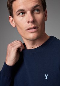 Maglione a maglia blu navy con collo tondo, polsini a coste e un piccolo logo di un cervo azzurro sul lato sinistro del petto. Tessuto morbido, vestibilità casual.