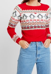 Röd tröja med vit och grå pingvinmönster, prydd med silver- och röda detaljer. Ribbstickade muddar och nederkant. Bärs med blå jeans.