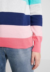 Randig tröja i turkos, grå, marinblå, rosa och korall. Stickad textur, ribbade ärmmuddar och fåll; figurnära silhuett. Bärs med denimjeans.