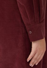 Burgundowa koszula z korduruyu z długimi rękawami, z guzikowym mankietem i pionowym prążkowanym wzorem. Zbliżenie na rękaw i dłoń.