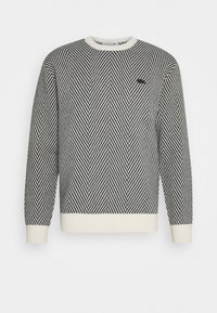 Svart och vit herringbone-mönstrad crewneck-tröja med vita muddar och nederkant, med en liten svart broderad logotyp på bröstet.