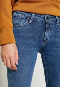 Blue denim jeans with a slim fit, featuring standard pockets, light whiskering, and a contrast orange stitching detail.