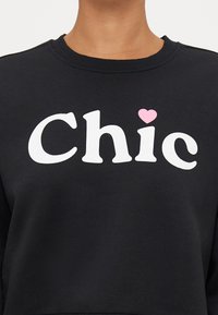 Μαύρη μπλούζα φόρμας με λευκό εκτυπωμένο κείμενο σε μπλοκ γράμματα που αναγράφει "Chic" με ροζ διακοσμητική καρδιά. Μαλακό ύφασμα, στρογγυλή λαιμόκοψη και ριμπ μανσέτες.