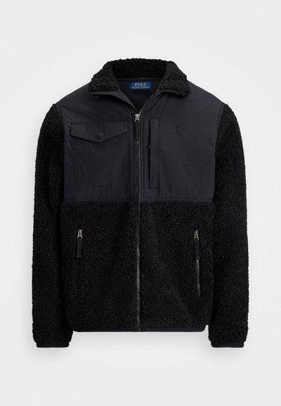 Mustmust muströvel, millel on tekstureeritud kangas, kaks esist zip-taskut, rinnaflap tasku ja tõusnud krae.