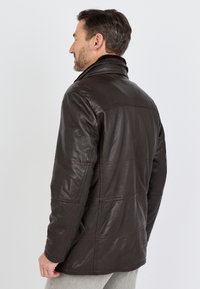 Man met kort donker haar en baard, die een donkerbruin leren jack draagt, kijkt naar links met de achterkant grotendeels zichtbaar tegen een effen achtergrond.