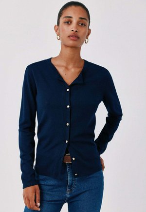 Donna che indossa un cardigan blu navy con bottoni e jeans blu, in piedi con una mano sul fianco contro uno sfondo chiaro e uniforme.
