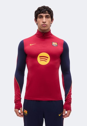 Mężczyzna noszący czerwoną i granatową sportową bluzę z długim rękawem oraz spodnie z logo FC Barcelona i Spotify, stojący przodem.