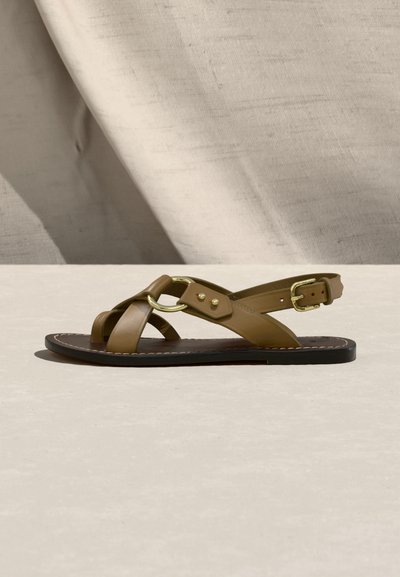 Brūnas ādas plakanās sandales ar krustojumiem siksniņām, zelta sprādzi un gredzena detaļu, attēlotas uz neitrālas bēšas virsmas un fona.