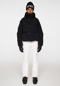 Svart pufferjacka med hög krage, stora framfickor och knäppning, tillsammans med vita byxor, svarta handskar och en svart mössa.