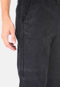 Pantaloni in velluto a coste blu scuro con texture a coste verticali, mano in una posizione naturale vicino alla tasca che mostra il dettaglio della cucitura laterale.