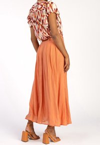 Gonna maxi plissettata arancione abbinata a una maglietta fantasia. L'outfit presenta maniche arricciate e sandali con tacco largo. Tessuto liscio e design fluido.