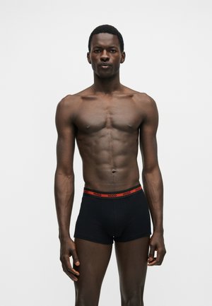 Atletyczny mężczyzna o ciemnej karnacji, ubrany w czarne bokserki z czerwonym paskiem HUGO, stojący prosto na jednolitym białym tle.