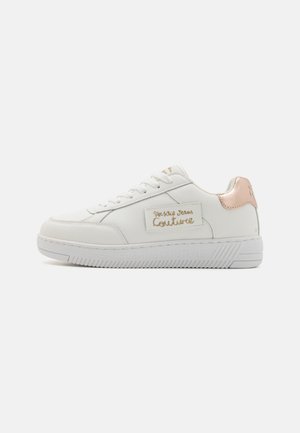 White sneaker with a textured leather upper, featuring a blush pink heel tab, gold logo, and white rubber sole with ribbed detailing.