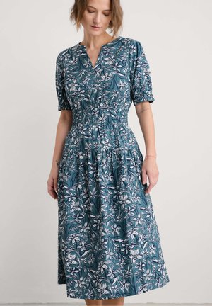 Teal mekko, jossa on lyhyet puffihihat, V-pääntie, kootut vyötäröt ja valuvan hameen, jossa on valkoisia kukka- ja lehtikuvioita.