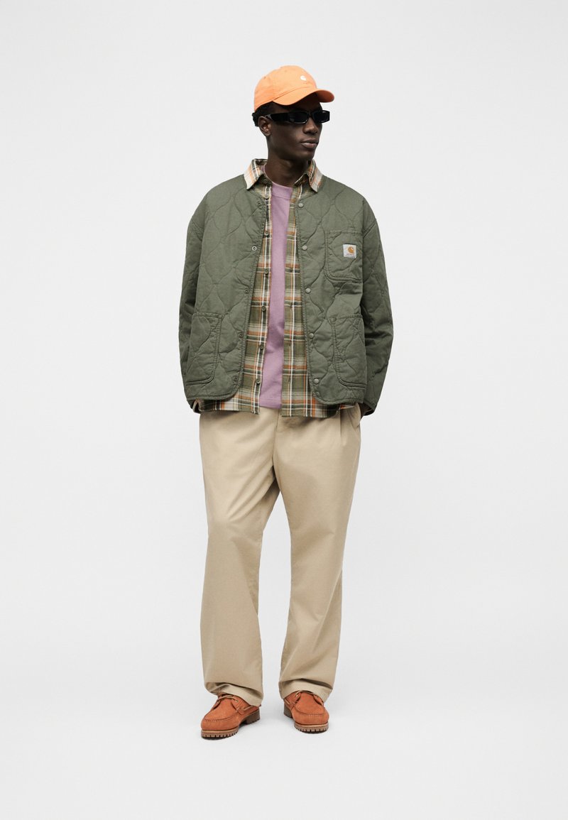 Homme portant une casquette orange, des lunettes de soleil noires, une veste matelassée verte, une chemise à carreaux, un T-shirt violet, un pantalon beige et des chaussures orange, debout sur un fond blanc.