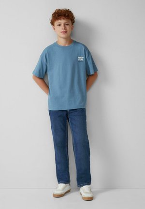Blaues Baumwoll-T-Shirt mit kurzen Ärmeln, mit dem Aufdruck "INNOVATIVE". Dazu dunkelblaue Jeans und weiße Turnschuhe mit beigefarbenen Sohlen.