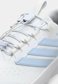 Zapatilla deportiva blanca con parte superior texturizada, cordones elásticos, franja de acento azul y lengüeta trasera reforzada para mayor soporte.