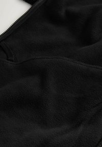 Černá fleece látka s měkkou texturou, vybavená zipovým uzávěrem a šitými detaily. Povrch se zdá být hladký s mírnými záhyby.