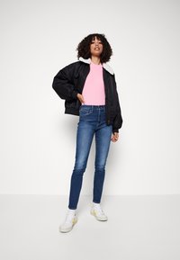 Czarna kurtka bombowca z białym kołnierzem z sztucznego futra, różowy prążkowany top, niebieskie dopasowane dżinsy i białe sneakersy z żółtymi akcentami.