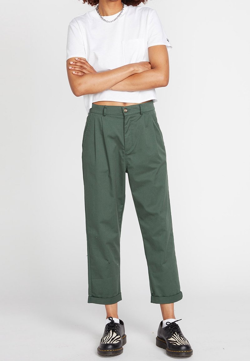 Grønne plisserede bukser lavet af bomuld, med lige ben og opføjede kanter. Kombinationen er med en hvid korteret t-shirt og sort-hvide sko.