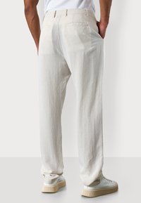 Model wearing light-colored trousers with back pockets, hands in pockets, paired with white sneakers, against a neutral background.