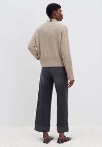 Beige gestrickter Pullover mit hohem Kragen, lockerer Passform und geripptem Saum, kombiniert mit dunkelgrauen weit geschnittenen Jeans, die an den Knöcheln umgeschlagen sind.