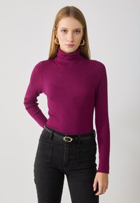 Tettskjorte med høy hals i ribbet tekstur, livlig magenta farge, paret med høy midje sorte jeans og et enkelt svart belte.