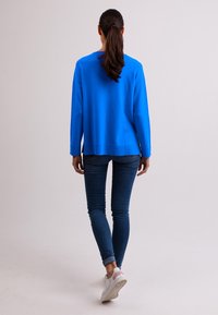 Blauer Pullover mit rundem Halsausschnitt und langen Ärmeln, kombiniert mit dunklen Jeans und weißen Sneakers. Glatte Textur, lockere Passform.