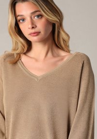 Beige, lichtgewicht trui met een V-hals en lange mouwen. De textuur is soepel met een subtiele glans, en de stof lijkt zacht.