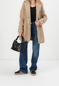 Měkký béžový kabát s kapucí, modré rozšířené džíny z denimu a černá kabelka s texturovaným vzorem. Černé tenisky s bílými pruhy doplňují vzhled.