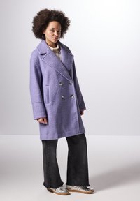 Cappotto viola doppiopetto con finitura testurizzata, ampi risvolti e tasche frontali, abbinato a jeans neri a zampa d'elefante e scarpe argento.