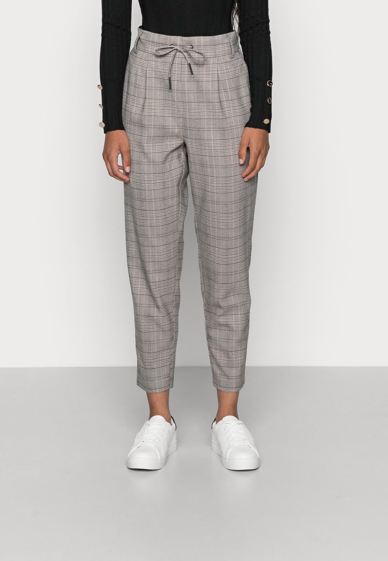 Femme portant un pantalon gris à carreaux fuselé avec une taille à cordon, un haut noir à manches longues avec des boutons dorés et des baskets blanches, debout sur un sol gris.