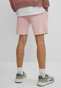 Homme portant un short rose clair, des chaussettes blanches et des sneakers gris, se tenant contre un fond gris uni, vu de dos.