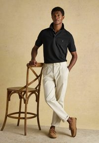 Homme en polo noir et pantalon beige appuyé sur une chaise en bois sur un fond beige uni.