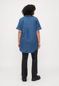Denim krekls ar īsām piedurknēm, apkakli un noapaļotu apakšmalu. Aizmugurējā kroka detaļa. Nēsāts ar melnām pieguļošām biksēm un masīviem melniem apaviem.
