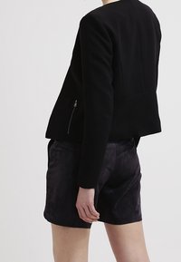 Veste noire sur mesure avec un col rond, des manches longues et des accents de zip latéraux, associée à un short en suède gris foncé.