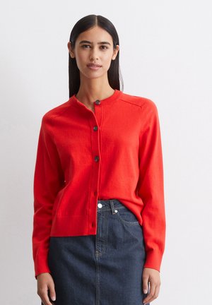 Rote Strickjacke mit rundem Halsausschnitt, fünfknöpfigem Verschluss und gerippten Bündchen. Kombiniert mit einem dunklen Jeansrock.