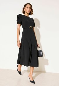 Crna haljina s napuhanih rukava, strukiranog struka, detaljem zlatne kopče i lepršavom suknjom. Uparena s crnim visokim potpeticama i malom torbicom.