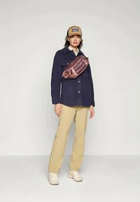 Tumesinise fliisjakiga, millel on tekstureeritud viimistlus, beežid püksid ja pruun müts. Kehast üle risti kulgev roosakas-pruuni värvi vöö kott logoväärsusega.