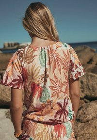 Pitkähiuksinen nainen pukeutuneena lyhythihaiseen kukkamekkoon, jossa on oransseja, vihreitä ja punaisia lehtiä, seisoo kivisellä rannalla kirkkaan taivaan alla.